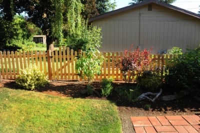 1006 Fencing Cedar Picket A dog eared cedar picket fence surrounded by bushes.